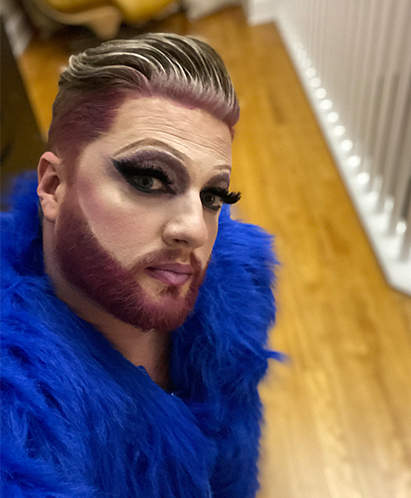 Man dressed in drag with makeup and tinted beard/false eyelashes.