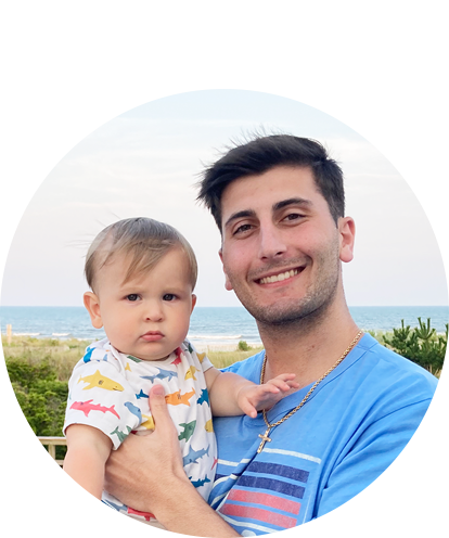 Daniel DeMarco fun photo (adult male posing holding young infant outdoors on sunny day with body of water in background).