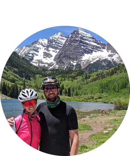 Leslie Riggs fun photo (adult female and adult male posing in bicycle helmets and sunglass with mountains and mountain lake in background).